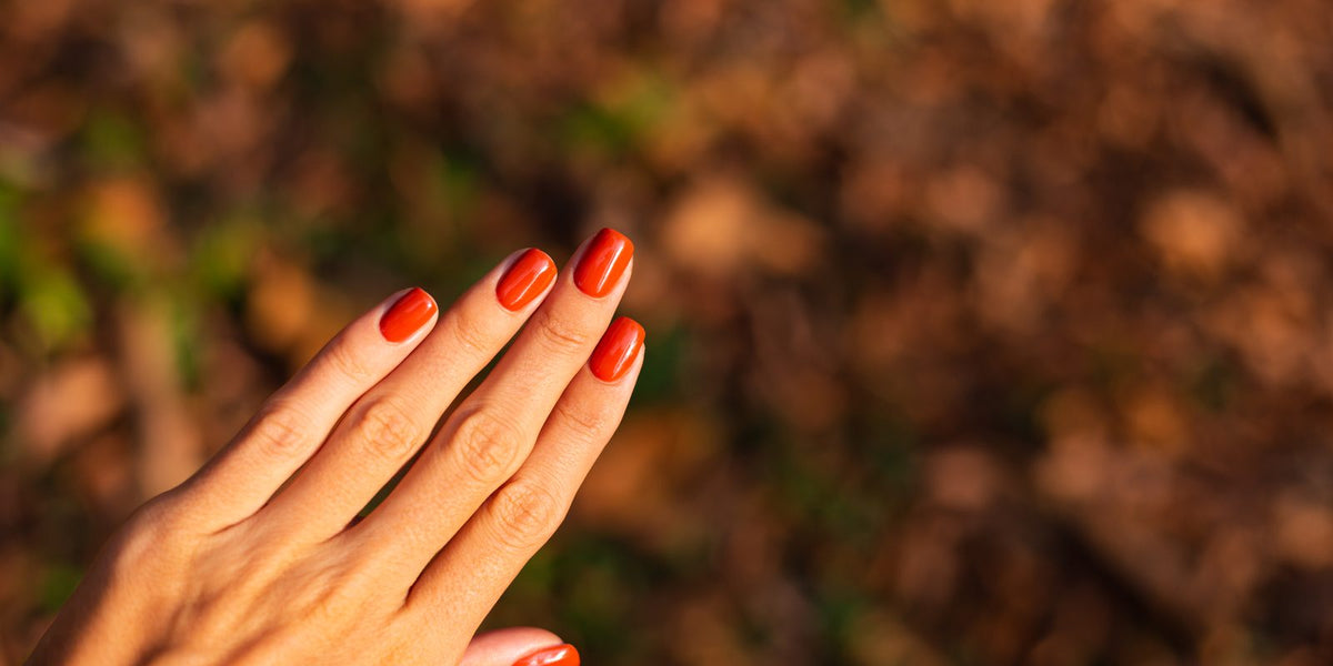 Orange-red manicure held over a blurred background of brown autumn leaves