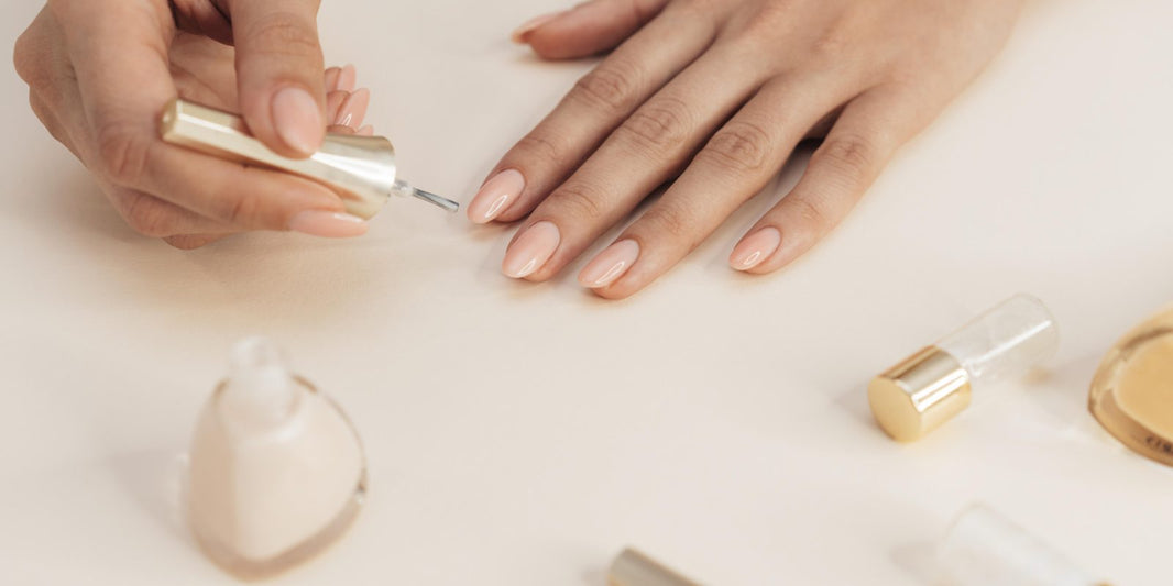 Hands with a fresh neutral gel manicure resting beside nail polish bottles on a beige surface