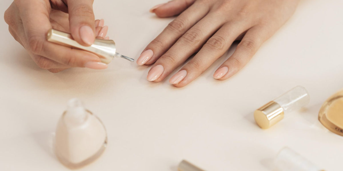 Hands with a fresh neutral gel manicure resting beside nail polish bottles on a beige surface
