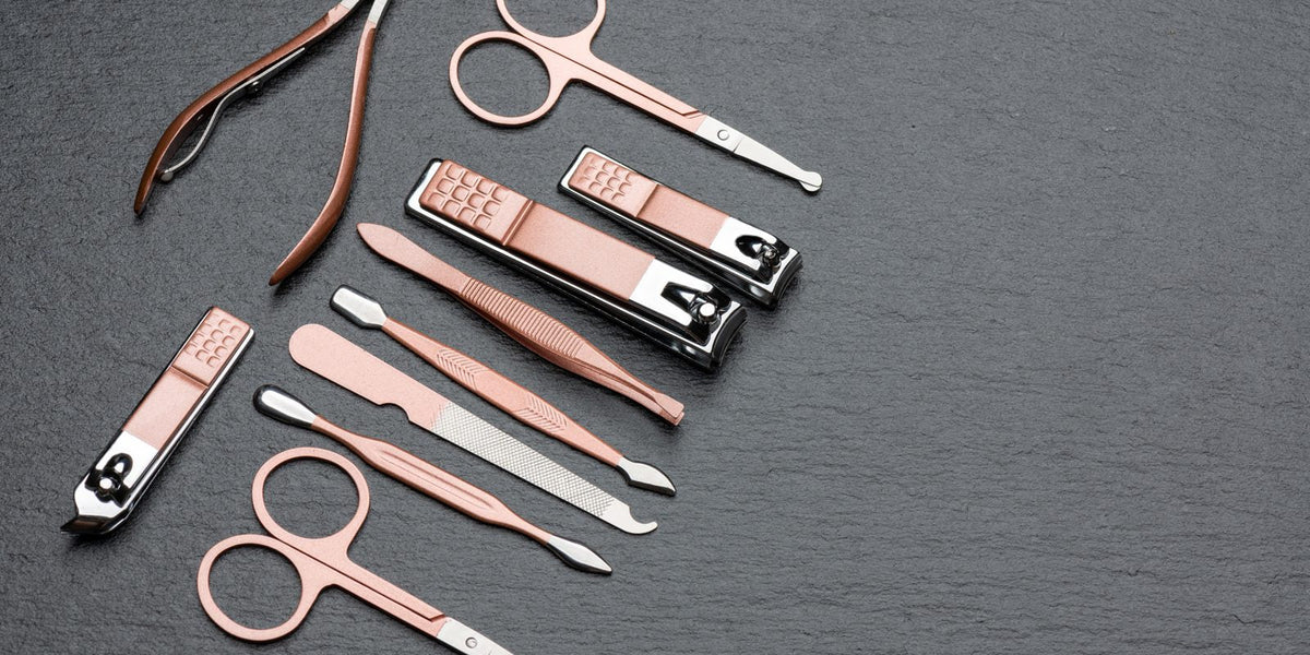 Rose gold nail clippers, a cuticle nipper, scissors, tweezers and a nail file on a dark grey stone surface